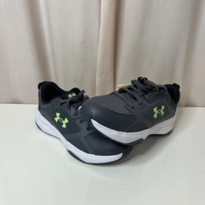 Under Armour skor - Snygga grå sneakers från Under Armour med limegrönt logotryck på sidan. Skorna har en vit, platt sula och är tillverkade i syntetmaterial med meshdetaljer för extra ventilation. Klassisk snörning och rund tå gör dem perfekta för träning eller chill vardagsstil.