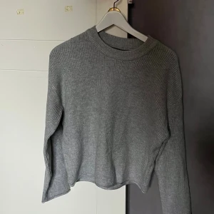 Grå ribbad stickad tröja - En stilren grå stickad tröja med ribbad struktur och rund hals. Tröjan har lång ärm och en något croppad passform som ger en modern vibe. Perfekt att matcha med jeans eller kjol för en chill look.