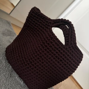 Brun handgjord stickad handväska - Unik brun handväska med grovstickad design och integrerade handtag. Väskan har en fyrkantig form och är tillverkad i mjukt stickat material som ger en cool och chunky vibe. Perfekt för dig som gillar personliga accessoarer med lite extra karaktär.