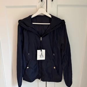 Mörkblå vindjacka från Moncler - Snygg mörkblå vindjacka från Moncler med huva och dragkedja framtill. Jackan har snörning vid huvan, två fickor med tryckknappar och lätt, tunn polyesterkänsla. Perfekt för dig som gillar stilrena och sportiga plagg med exklusiv känsla.