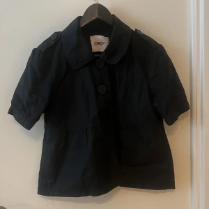 Svart kort trenchcoat från ONLY - Snygg svart trenchcoat från ONLY med korta ärmar och stora knappar framtill. Jackan har klassisk krage, axelklaffar med knapp och två fickor. Perfekt för dig som vill ha en stilren och enkel look med lite edge.