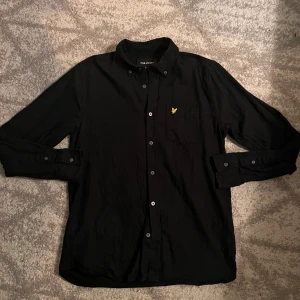 Svart skjorta från Lyle & Scott, M - Svart skjorta från Lyle & Scott i storlek M, tillverkad i mjuk bomull. Klassisk krage, långa ärmar och knäppning framtill med svarta knappar. På bröstet finns den ikoniska gula Lyle & Scott-loggan broderad. Perfekt för en clean och stilren look.Pris går att diskutera vid snabb affär!!