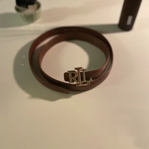 Brunt skärp från Ralph Lauren - Snyggt brunt bälte från Ralph Lauren med en stilren RLL-spänne i guld. Bältet är gjort i slätt skinn och har klassisk form med fina sömmar längs kanterna. Perfekt för att lyfta din outfit med en exklusiv touch och ikonisk logga framtill.
