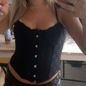 Svart spetskorsett med knäppning - Säljer en svart korsett med spetsdetaljer och snygg knäppning framtill. Modellen är croppad och har smala axelband. Perfekt för dig som vill ha en edgy och trendig look. Passar till både jeans och kjol.