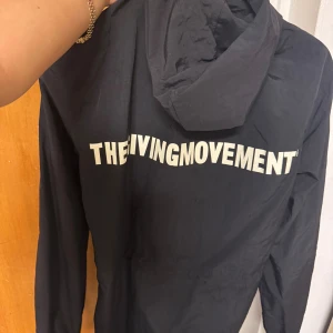 Svart vindjacka från The Living Movement - Svart vindjacka med huva från The Living Movement. Jackan har vit logga tryckt på ryggen, dragkedja framtill och justerbar dragsko i huvan. Lätt och smidig modell med långa ärmar och elastiska muddar. Perfekt för blåsiga dagar. Ny pris 1600kr