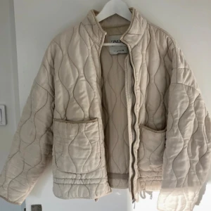 Beige quiltad jacka från Zara - Quiltad beige jacka från Zara med oversized passform, stora fickor framtill och dragsko i nederkant. Jackan har vågformat mönster i sömmarna och stängs med dragkedja. Perfekt för lager-på-lager och chill stil.