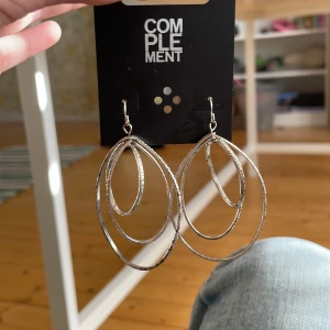 Stora silvriga örhängen från Complement - Säljer ett par stora, statement örhängen i silverfärgad metall från Complement. Örhängena har flera ovala ringar i olika storlekar som hänger i lager och ger en cool, modern look. Perfekta för dig som vill sticka ut och addera lite edge till din outfit.