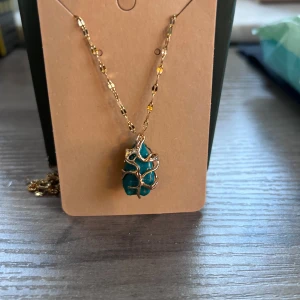 Halsband med grön sten och guldkedja - Unikt halsband med en grön sten infattad i ett dekorativt guldfärgat nät. Kedjan är tunn och glittrig med små detaljer som fångar ljuset. Hänget har en organisk, naturinspirerad form som verkligen sticker ut.