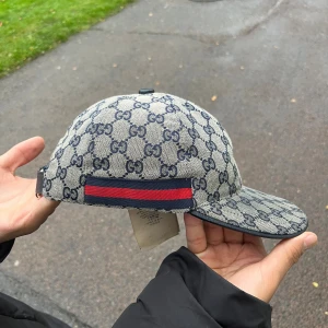  Äkta Gucci keps med GG-mönster och röd-blå detalj - Snygg keps från Gucci med klassiskt GG-mönster i mörkblått på beige botten. Kepsen har en böjd skärm och justerbar rem bak i mörkblått skinn. Sidorna pryds av en röd och marinblå rand för extra lyxig vibe. Perfekt för dig som vill sticka ut med designerstil.