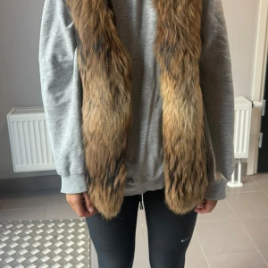 Woolrich - Brun dunväst med stor päls  - Säljer en mörkbrun dunväst med stor fluffig päls runt huvan. Västen har en lång, figurnära passform och praktiska fickor med dragkedja på sidorna. Pälsen är brun med svarta inslag och ger en lyxig känsla. Perfekt för kalla dagar när du vill ha både stil och värme.