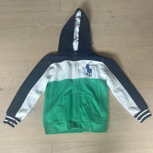  Hoodie från Polo Ralph Lauren - Snygg original hoodie från Polo Ralph Lauren i färgblock med marinblå, vit och grön. Tröjan har dragkedja, huva, två fickor fram och klassisk Polo-logga på bröstet. Ribbade muddar med ränder på ärmsluten ger en sportig känsla. Perfekt för dig som gillar streetstyle. Storlek 7år