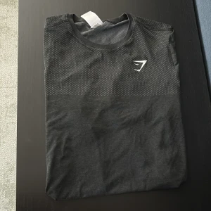 Svart Gymshark t-shirt med prickigt mönster - Snygg svart t-shirt från Gymshark i storlek M. Övre delen har ett diskret prickigt mönster och en vit Gymshark-logga på bröstet. Tillverkad i en blandning av nylon och polyester, perfekt för träning. Klassisk rund hals och korta ärmar. Använd 1 gång 
