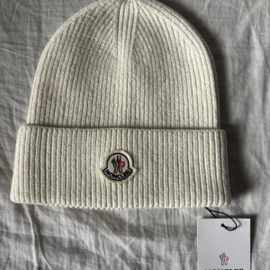 Vit ribbad Moncler mössa - Snygg vit ribbstickad mössa från Moncler med klassisk uppvikt kant och broderad logga framtill. Mössan är tillverkad i mjuk ull och har en clean, stilren look som passar perfekt till kalla dagar. Taggen sitter kvar för extra exklusiv känsla.