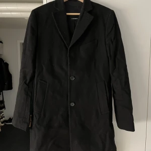 Svart rock från J.Lindeberg, strl 46 - Stilren svart rock från J.Lindeberg med klassisk krage, två knappar framtill och fyra dekorativa knappar vid ärmslutet. Kavajen har raka fickor och en bröstficka, perfekt för dig som gillar en clean och modern look. Materialet är mjukt och känns lyxigt.