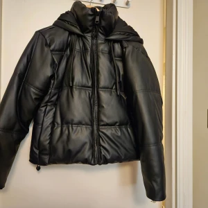 Svart pufferjacka i skinnimitation Zara XS - Säljer en svart pufferjacka från Zara i skinnimitation, storlek XS. Jackan har huva, dragkedja framtill och dragsko nertill. Den är quiltad och har en modern, boxig passform som är perfekt för kalla dagar.