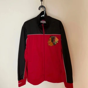 Chicago Blackhawks   Mitchell & Ness - Snygg röd och svart tröja från Mitchell & Ness med broderad Chicago Blackhawks-logga på bröstet. Tröjan har hel dragkedja framtill, ståkrage och vita detaljer längs bröst och ärmar. Perfekt för dig som gillar sportig stil och hockey.