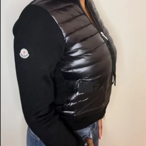 Moncler cardigan  - Snygg svart dunväst från Moncler med quiltad framsida och två fickor med knappdetalj. Västen har dragkedja framtill och Moncler-logga på ärmen. Perfekt för lager-på-lager och ger en sportig vibe.