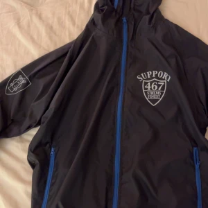 Svart vindjacka Support 467 Sthlms Finest - Svart vindjacka med blå dragkedjor och tryck på bröstet och ryggen. Jackan har huva, två fickor med dragkedja och tryck på ena ärmen. Perfekt för hösten och våren