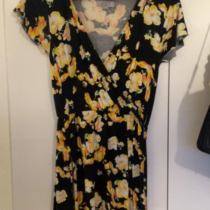 Svart blommig omlottklänning - Svart omlottklänning med v-ringning och korta ärmar. Klänningen har ett färgglatt blommönster i gult och vitt, markerad midja och faller lätt. Perfekt för sommarens alla tillfällen.