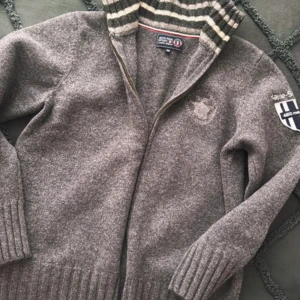 Grå stickad kofta Jean Paul  - Säljer en grå kofta från Jean Paul Koftan har dragkedja, ribbade muddar och krage med gråvita ränder. Broderad logga på bröstet och patch på ena ärmen. Perfekt för kyliga dagar och snygg till jeans.