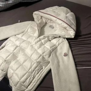 Vit quiltad dunjacka från Moncler - Snygg vit dunjacka från Moncler med quiltad front och huva. Ärmarna är stickade i mjuk ullmix och jackan har dragkedja framtill. Klassisk Moncler-logga på ärmen och diskreta röda detaljer på huvan. Perfekt för kyliga dagar när du vill vara både varm och stilren.