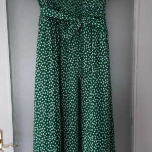 Grön blommig midiklänning med knytband - Säljer en grön midiklänning med vita små blommor och smock upptill. Klänningen har ett matchande knytband i midjan och är tillverkad i ett lätt, mjukt tyg som känns skönt mot huden. Perfekt för dig som gillar färg och mönster!