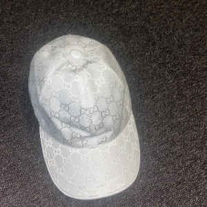 Vit Gucci keps med GG-mönster - Snygg vit keps från Gucci med det klassiska GG-mönstret över hela kepsen. Tillverkad i 100% bomull och har en böjd skärm. Insidan är brun med Gucci-logga på svettbandet. Perfekt för dig som vill ha en lyxig och clean look.