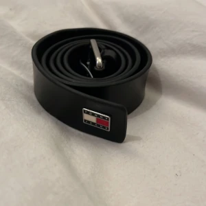 Svart skärp från Tommy Jeans - Snyggt svart bälte från Tommy Jeans med klassisk metallspänne och logotyp i silver, rött och vitt på änden. Bältet är tillverkat i slätt skinn och har en stilren, rak form som passar till jeans och andra outfits. Perfekt accessoar för att lyfta din look.