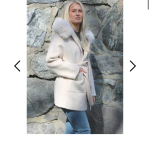 Fur Stockholm Alicia Coat off-white med pälskrage - Snygg off-white kappa från fur Stockholm med stor fluffig pälskrage och knytskärp i midjan. Jackan har raka ärmar, två fickor och en stilren, lite oversized passform. Perfekt för dig som vill ha en trendig och mysig look under kallare dagar.