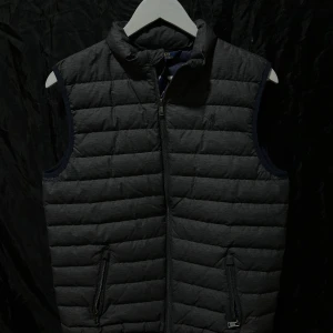 Grå dunväst från Polo Ralph Lauren - Snygg grå dunväst från Polo Ralph Lauren med quiltad design och ståkrage. Västen har två praktiska fickor med dragkedja och en mörkblå insida. Perfekt att slänga över en hoodie för extra värme. Stängs med dragkedja framtill. Det står storlek XL/TG (18-20) förstår inte riktigt vad detta betyder men skulle säga att den är i vanlig S storlek. Hör av er vid frågor och pris kan diskuteras vid snabb affär!