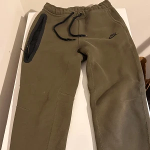 Olivgröna mjukisbyxor från Nike - Snygga olivgröna mjukisbyxor från Nike med svart logga på benet. Byxorna har en bred resår i midjan med svarta snören och en stor ficka med dragkedja på sidan. Perfekta för chill eller träning, tillverkade i mjukt bomullsmaterial.