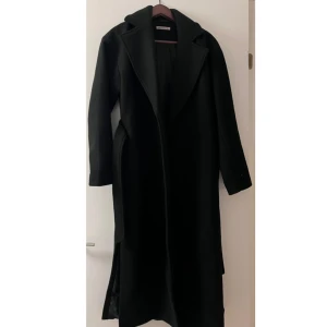 Svart lång kappa med bälte - Säljer min elegant utformade, svarta kappa från Anna Field. Plagget är i utmärkt skick och har endast använts vid ett fåtal tillfällen. Kappan har en tidlös och sofistikerad design med djup krage, rak passform och diskret slits baktill, vilket skapar en stilren och modern silhuett.  Ett klassiskt ytterplagg som passar lika väl till formella tillfällen som till en mer avslappnad vardagsstil. Köpte den för 1699kr men säljer den för 1200kr priset kan diskuteras
