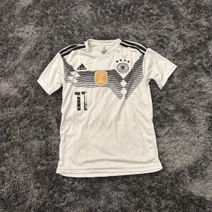 Tysklands Reus #11 fotbollströja Adidas - Säljer en vit Tysklands landslagströja från Adidas med Reus och nummer 11 på ryggen. Tröjan har svarta ränder och detaljer, FIFA World Champions-märke på bröstet och broderat landslagsemblem. Kortärmad och tillverkad i lätt polyester, perfekt för match eller träning.
