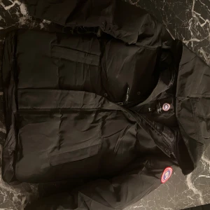 Svart dunjacka från Canada Goose - Säljer en svart dunjacka från Canada Goose med huva och klassisk logga på ärmen. Använt i bara en vecka. Jackan har flera fickor framtill, dragkedja och quiltad design. Perfekt för kalla dagar och riktigt snygg streetstil.