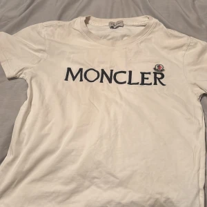 Vit Moncler t-shirt med logga - Vit t-shirt från Moncler med stor broderad logga i svart på bröstet och klassisk rund hals. T-shirten har korta ärmar och är gjord i mjuk bomull. Snyggt broderad Moncler-emblem i svart och rött vid loggan. Perfekt för dig som gillar exklusiva streetwear-vibbar.