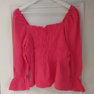 Rosa blus med puffärmar, stl L - Supersöt rosa blus med smockad framsida och vida puffärmar som avslutas med volang. Blusen har en fyrkantig ringning och är tillverkad i ett lätt tyg som känns luftigt. Perfekt för dig som vill ha en färgklick i garderoben!