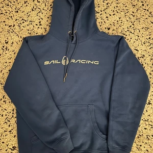 Blå set från Sail Racing - Blå set från Sail Racing med snyggt tryck på bröstet och justerbar snörning i huvan. Tröjan har en klassisk känguruficka framtill och ribbade muddar vid ärmslut och nederkant. Perfekt för chill dagar och streetwear-stil.