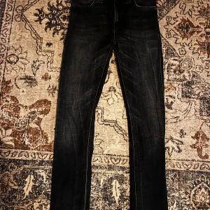 Svarta jeans från Nudie Jeans Co 26/32 - Snygga svarta jeans från Nudie Jeans Co i storlek 26/32. Jeansen har klassisk femficksdesign, orangea detaljer och en mörk tvätt med lätt slitning. Passformen är rak och materialet är mjukt denim. Perfekta för dig som gillar stilrena och tidlösa jeans.