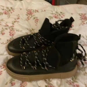 Svarta boots med snörning och mockadetaljer - Säljer ett par svarta boots med grov beige sula och snörning med svarta och vita skosnören. Ovandelen är i skinn och mocka, med högt skaft och rund tå. Insidan har mjuk mocka för extra komfort. Perfekt för dig som gillar chunky stil och vill ha något som sticker ut.