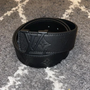 L-bälte  - L–bälte i svart med tonat monogram och svart spänne. Stilren och diskret design. Passar både herr och dam. Storlek 110 cm