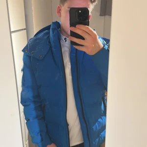 Blå pufferjacka från Amicci  - Säljer en blå pufferjacka från Amicci i storlek M. Jackan har huva med knappar och dragsko, hel dragkedja framtill och fickor med dragkedja. Materialet är syntet och jackan är perfekt för kalla dagar. Snygg och modern look. Köpt från Storbritannien, UK 🇬🇧 