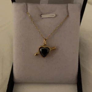 Halsband med svart hjärthänge och pil guld  - Guldfärgat halsband med ett svart hjärta som hänge, dekorerat med en pil genom hjärtat. Kedjan är tunn och elegant, och hänget har en cool, modern vibe med sin kontrast mellan guld och svart. Perfekt accessoar för dig som gillar unika detaljer. Rostfri 