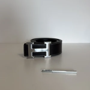 Svart skärp med H-spänne från H - Snyggt svart bälte från H med ett stort, silvrigt H-format spänne. Bältet är gjort i präglat skinn med vita sömmar längs kanterna. Perfekt accessoar för att lyfta din outfit med en lyxig touch.