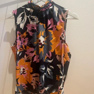 Blommig ärmlös blus med knyt - Säljer en färgstark blommig blus med högt hals och knytdetalj framtill. Blusen är ärmlös och har stora blommor i orange, rosa, grått och vitt mot en mörk bakgrund. Materialet är mjukt och har ett lätt glansigt utseende, perfekt för sommarens alla tillfällen.