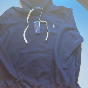 Marinblå hoodie från Polo Ralph Lauren - Snygg marinblå hoodie från Polo Ralph Lauren med klassisk broderad logga på bröstet. Tröjan har huva med vita snören och en stor magficka framtill. Tillverkad i mjuk bomull, perfekt för chill dagar eller när du vill ha en clean look.