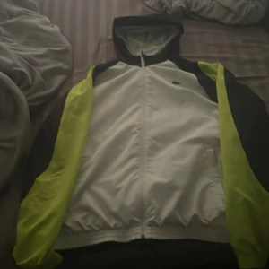 Lacoste Sport vit och gul vindjacka - Säljer en snygg vindjacka från Lacoste Sport i vitt med svarta och neongula detaljer. Jackan har huva, hel dragkedja och klassisk Lacoste-logga på bröstet. Perfekt för dig som vill ha en sportig och fräsch look.