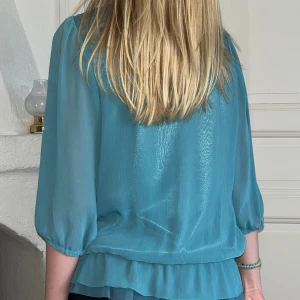 Turkos blus med spets och volang - Säljer en turkos blus i transparent, glansigt tyg med trekvartsärmar och volang nertill. Framsidan har fina veck och spetsdetaljer upptill vid axlarna. Blusen har en lös passform och är perfekt för dig som gillar romantiska detaljer.