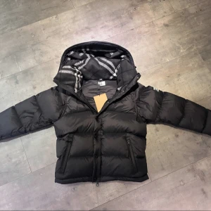 Burberry Puffer - Svart dunjacka från Burberry med klassisk Säljer en elegant och mångsidig Burberry-jacka i svart, i nyskick. Dunfodrad för värme och komfort med det klassiska rutmönstret i huvan som ger en exklusiv touch. Praktiska dragkedjefickor, avtagbar huva och diskret logopatch på ärmen. Passar M/L. Jackan är i princip som ny och utan defekter. En lyxig kombination av stil och funktion – perfekt för både vardag och tillfällen då du vill klä upp dig.  📦Snabb leverans - skickas samma dag📌
