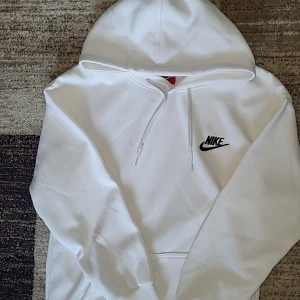 Vit hoodie från Nike med logga - Vit hoodie från Nike med svart broderad logga på bröstet. Tröjan har huva med snörning och ribbade muddar vid ärmslut och nederkant. Tillverkad i mjuk bomullsmix, perfekt för chill eller streetwear. Klassisk och clean design med skönt material.