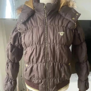 Brun pufferjacka med fuskpäls från JLO - Snygg brun pufferjacka från JLO med guldig dragkedja och detaljer. Jackan har avtagbar huva med fuskpäls, ribbade muddar och quiltad design. Perfekt för kalla dagar med sin varma och fluffiga känsla. Guldiga detaljer ger jackan en extra touch. Oanvänd 